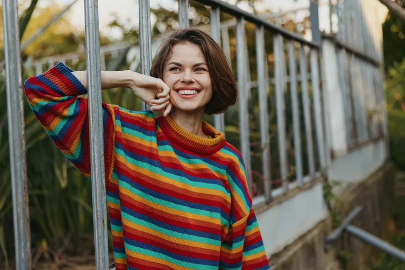 Smiling woman and woman in a colorful striped sweater leaning on a greenhouse frame outdoors; young adult with short brown hair in a casual outfit, joyful lifestyle portrait and relaxed mood.