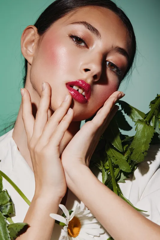 Close-up portrait of a fashionable woman with glossy red lipstick and dewy skin, holding green leafy plants and flowers. Concept for beauty creative fashion editorial, natural tones.