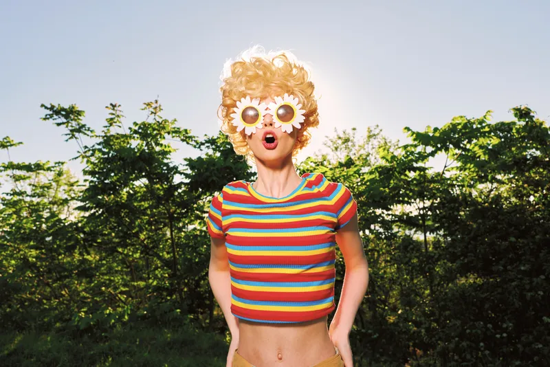 Playful young woman wearing a bright striped crop top and flower mask stands in a sunlit garden, her mouth open in a surprised, joyful expression as she enjoys a whimsical moment outdoors.