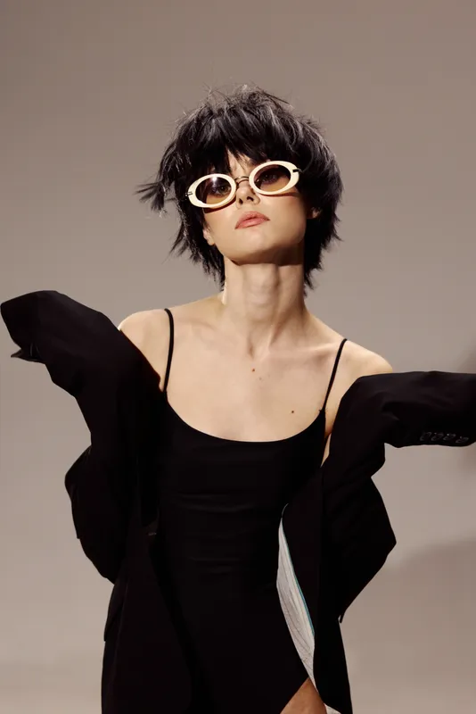 Stylish woman with short hair wearing white sunglasses and black dress posing confidently in studio lighting.