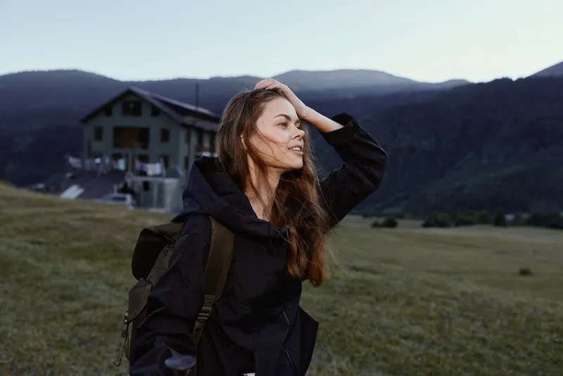 A young woman with a backpack stands in a grassy field, gazing toward distant mountains with a thoughtful smile, capturing a serene outdoor moment.