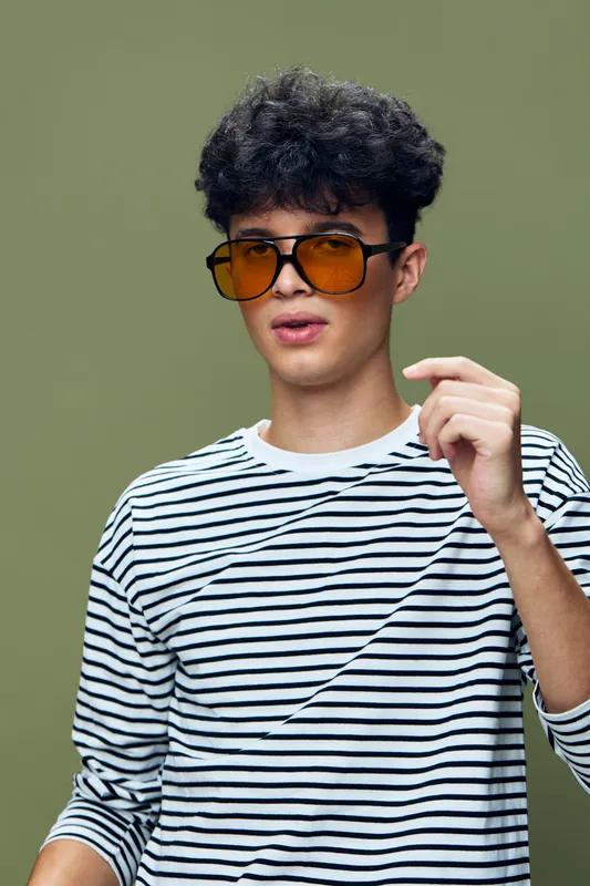 young man wearing striped shirt and large tinted sunglasses posing indoors with curly hair and neutral green background, casual style and cool expression