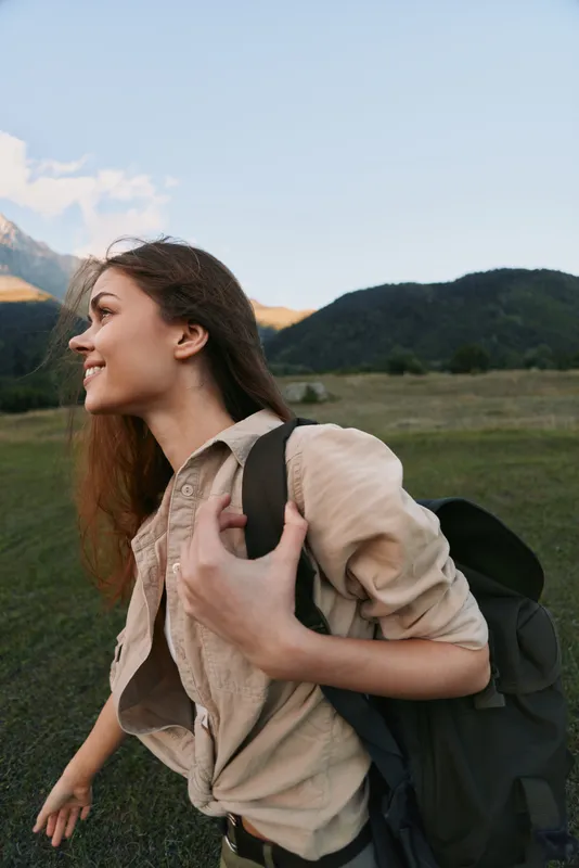 A young woman with a backpack hikes through a grassy meadow with distant mountains, smiling at the peaceful landscape during golden hour.