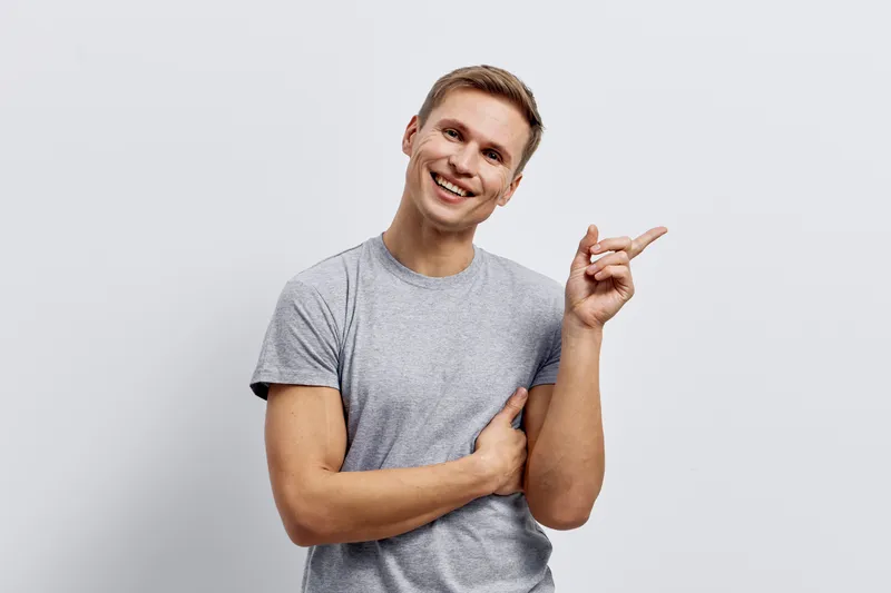 Smiling young man with short hair in casual gray t-shirt gestures with finger and looks at camera standing against plain white background cheerful and confident lifestyle concept