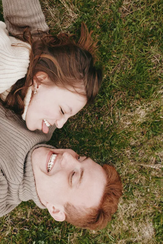 A young couple with red and brunette hair lies on green grass smiling and looking at each other enjoying a relaxed lifestyle moment outdoors.