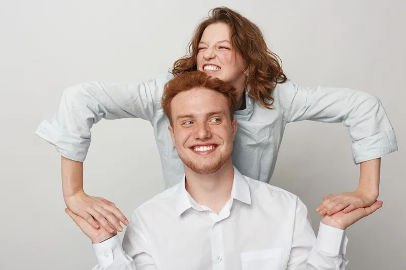 Playful young woman with curly hair leans on smiling redhead man in white shirts against light background. Concept of friendship, happiness, youth, and casual lifestyle together.