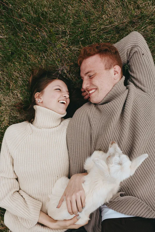 A young couple in cozy sweaters lies on the grass laughing together while holding a small white baby goat, enjoying a relaxed lifestyle moment outdoors in cool weather.