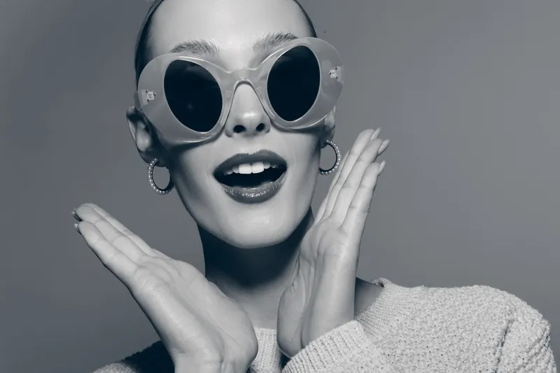 Surprised young white woman with fair skin wearing round sunglasses and hoop earrings expresses excitement with open mouth and hands near face. Studio portrait.