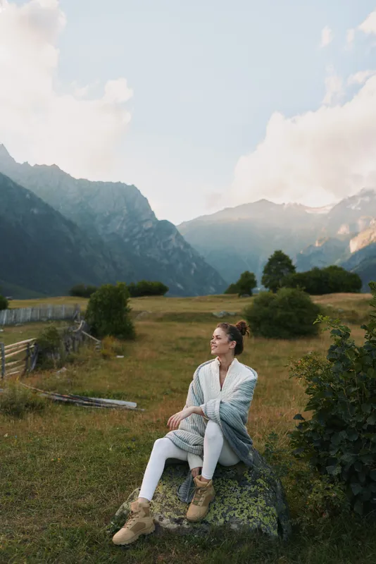 A woman sits on a rock in a meadow beneath mountains, wearing a light shawl and boots, enjoying outdoor life. A lifestyle concept focused on calm moments, nature, and relaxed fashion.