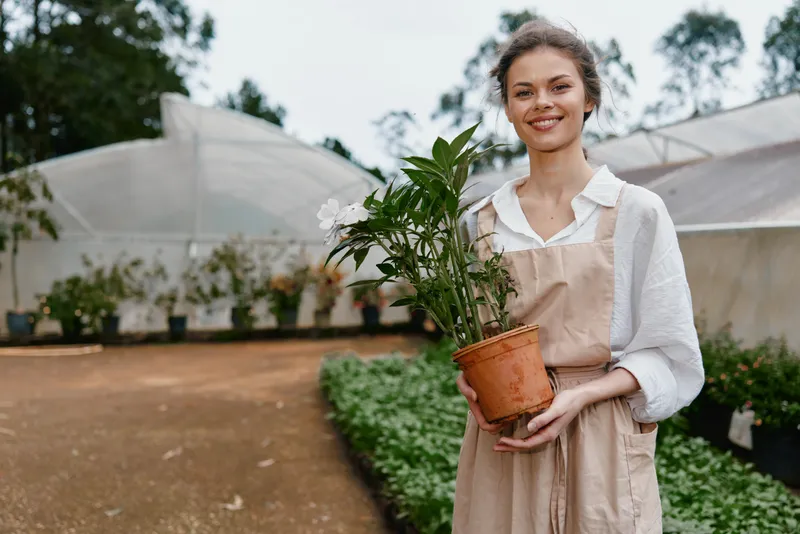 Young woman in an apron smiling while holding a potted plant, standing in a greenhouse with greenery in the background