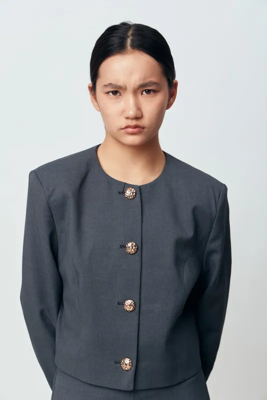 Young woman with serious expression wearing dark gray formal jacket standing isolated on white background, looking at camera with crossed arms and firm posture.