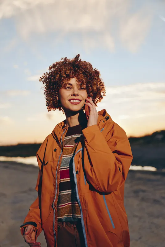 Smiling woman with curly hair wearing a tan jacket outdoors at sunset, walking on a road, warm lifestyle moment, soft golden light, autumn mood, natural portrait, in fresh air, during travel