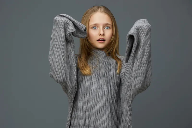 Young girl in oversized gray sweater expressing surprise with hands raised, against a neutral background, perfect for fashion and seasonal concepts