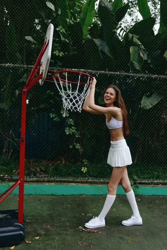 Cheerful young woman in sporty attire posing playfully near a basketball hoop, exuding joy and energy on a sunny day in a lush green setting