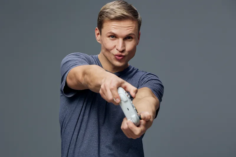 Playful young man with short light brown hair wearing casual blue t-shirt, expressing fun and engaging attitude while holding and pointing a game controller towards camera, isolated on gray background