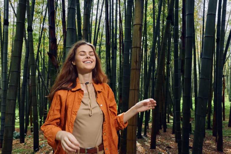 Woman in bamboo forest smiling with eyes closed, happy portrait in nature wearing an orange jacket, peaceful outdoor scene of relaxation and serene sunlight on trunks.