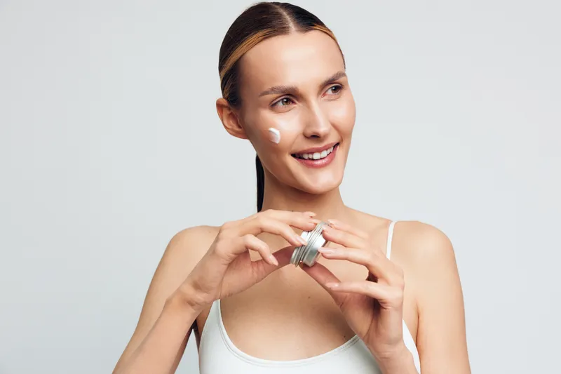 Woman in white top applies facial serum near chin smiling in studio light, focused skincare routine for beauty self care and hydration, clean minimal background, morning wellness gesture