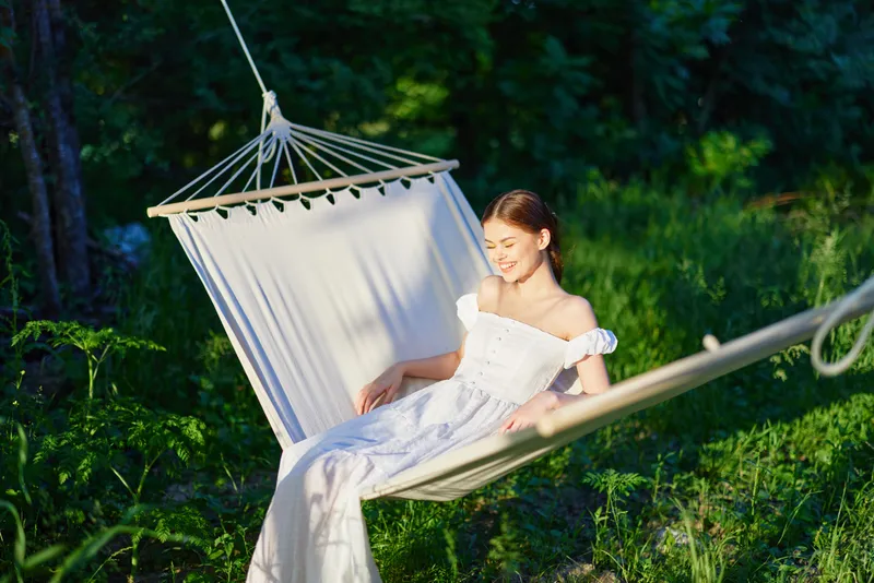 Young woman in a white dress relaxing in a hammock surrounded by greenery, showcasing a calm and joyful emotion in a summer setting