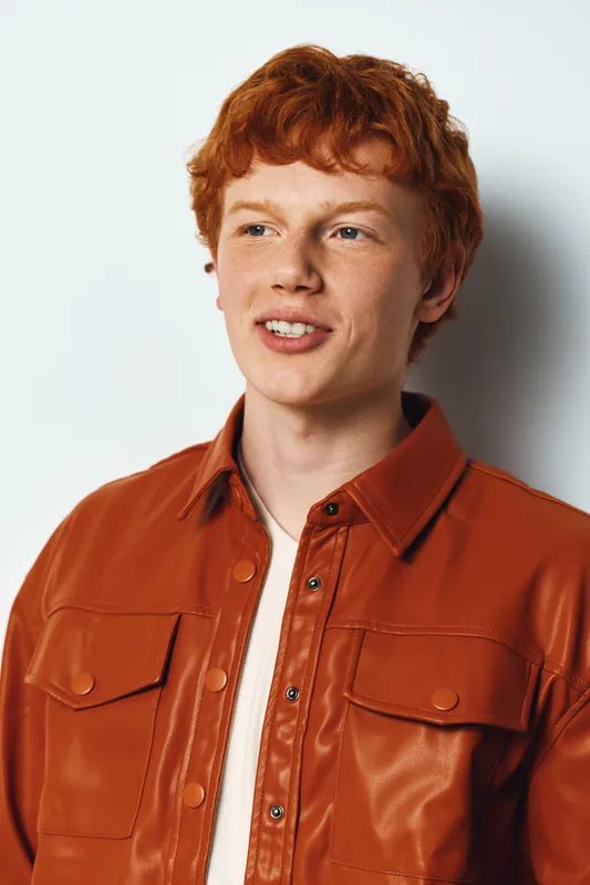 Young man with red hair and freckles wearing a brown leather jacket smiles looking away with white background. Casual style, portrait and confident expression for lifestyle concept.