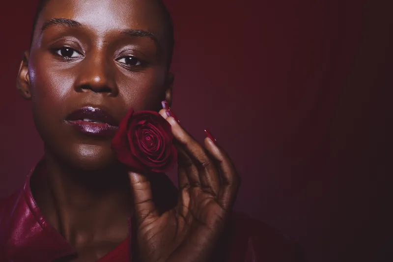 Close-up of a dark skinned African woman holding a red rose near her face. The studio portrait features glossy lips and nails, deep red tones, and elegant mood with soft lighting.