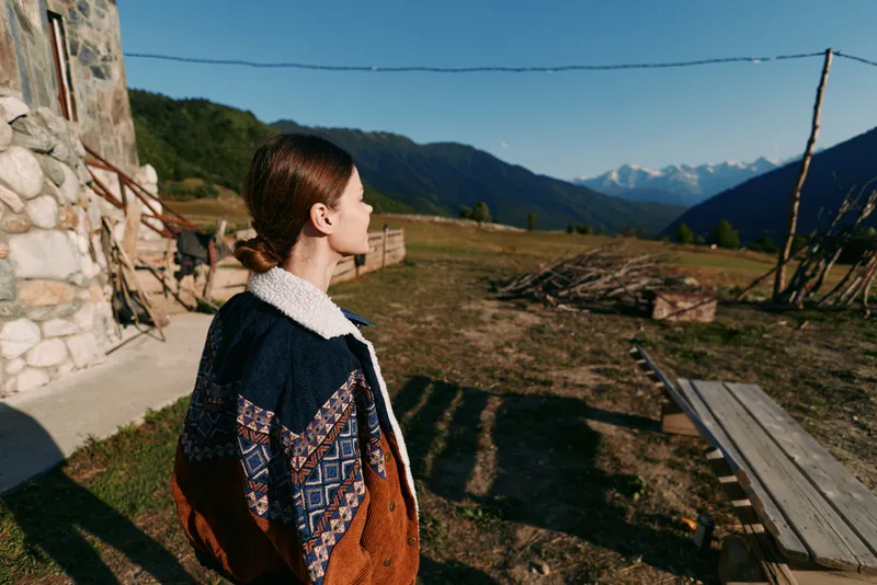 Woman in patterned jacket stands in rural countryside looking at mountain landscape, outdoors scene with wooden fence, stone building and clear sky for travel and nature.