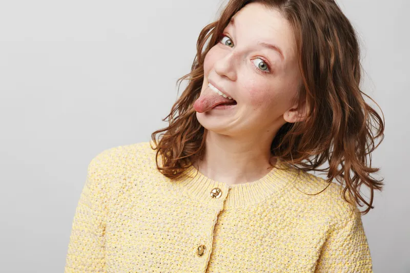 Playful woman with curly hair wearing yellow sweater sticks out tongue and smiles. Studio portrait on light background. Concept of fun, humor, and carefree attitude in casual fashion.