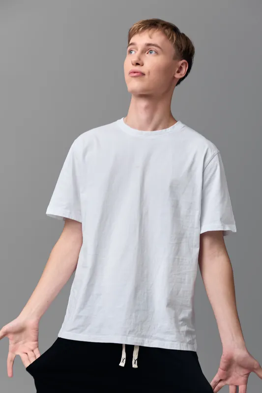 Young man in a white tee with open gesture stands in a neutral studio, conveying casual confidence, simple style, and effortless charm.