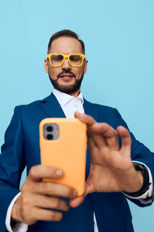 A professional man in a blue suit and yellow sunglasses takes a confident pose with an orange smartphone against an isolated colorful background, evoking bold energy and friendly mood.