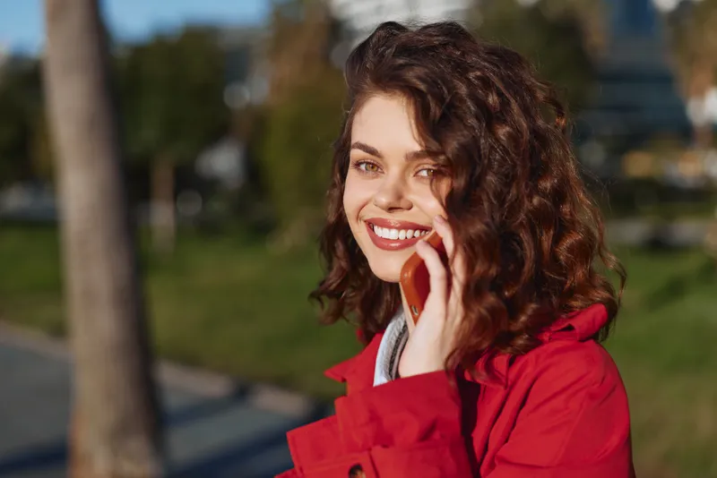 A joyful woman in a red jacket chats on a phone outdoors, smiling at the camera in a sunlit park. A candid portrait conveying warmth, connection, and everyday happiness.