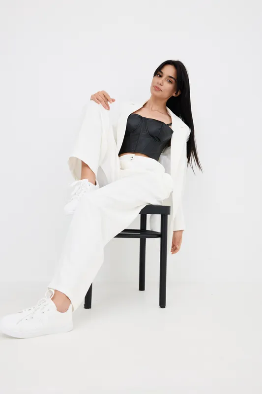 Stylish woman sitting on a black chair wearing white pants, white jacket, black corset top, and white sneakers against a clean white background, looking relaxed.