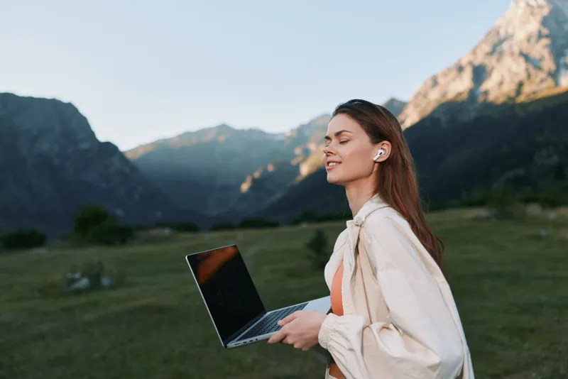 A focused professional woman stands outdoors in a scenic mountain landscape, holding a laptop. She smiles slightly, enjoying a productive moment in nature.