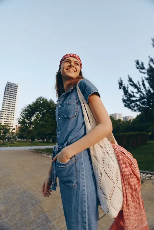 Lifestyle woman wearing modern boho-western denim street style outfit with a red bandana and crochet bag smiling outdoors in a film color setting under clear sky.