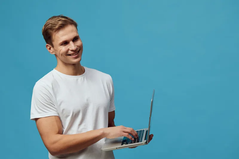 smiling man holding laptop in white t-shirt working digital nomad remote job on blue background studio technology concept