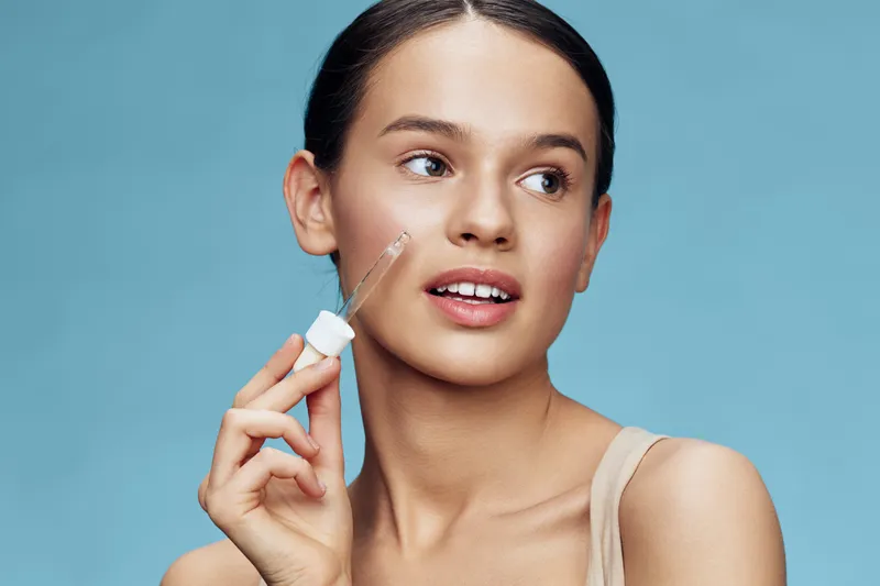 Young woman applying skincare serum on her face against a light blue background, showcasing natural beauty and healthy skin
