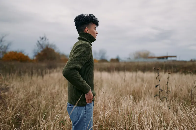 Side profile of a young man standing in tall autumn grass, candid pose and natural expression conveying authenticity and quiet confidence in a raw, credible outdoor portrait.