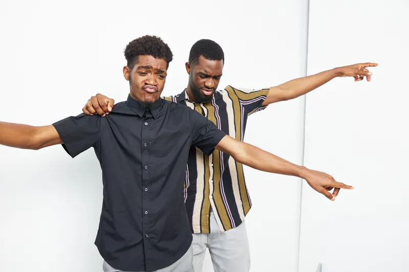 Two black men having fun together, expressing emotions while pointing in different directions. Their playful interaction showcases friendship and joy in a light atmosphere.