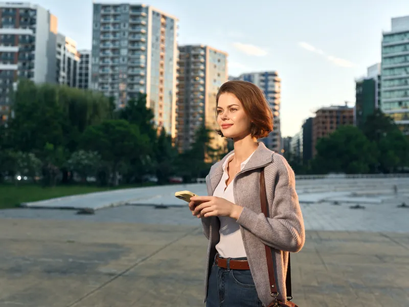 A cheerful business professional woman stands outdoors in an urban park, texting on her phone with a confident smile, while modern buildings rise in the background, conveying energy and focus.