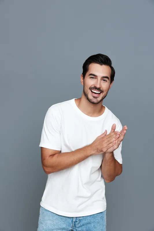 Smiling young man in a casual white t-shirt clapping hands, looking at the camera against a plain grey background. Expressing joy and enthusiasm in a vibrant portrait.