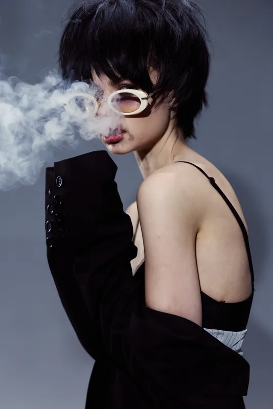 Woman with short hair smoking, wearing sunglasses and black clothing in a moody setting with dark background.