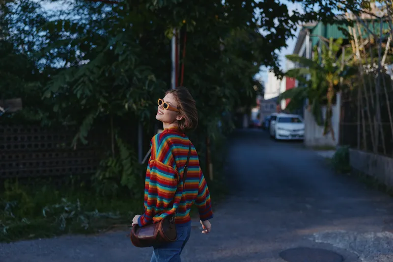 Woman in a colorful striped sweater and sunglasses walking a quiet street at evening, smiling and looking up in a casual outfit with small leather bag for relaxed travel lifestyle.