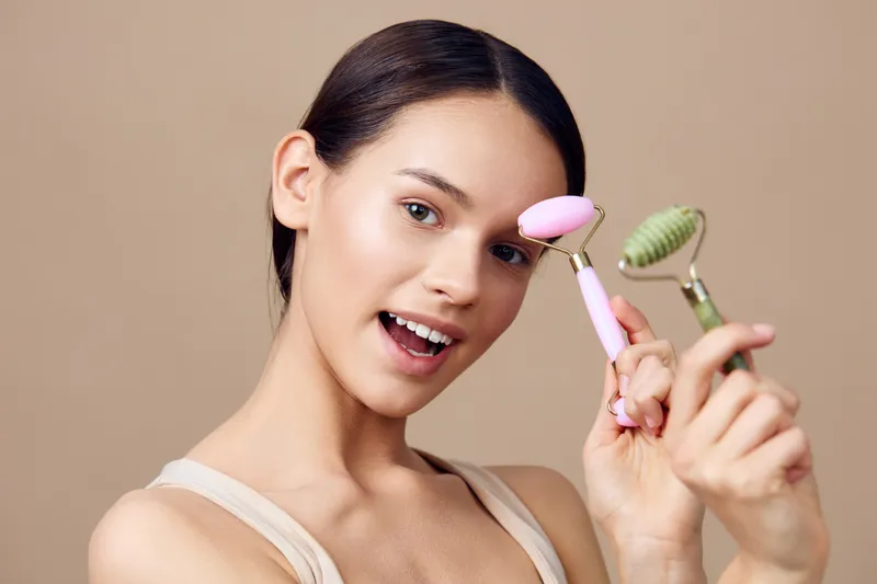 Young woman joyfully holding facial rollers in a neutral background, showcasing a beauty routine focused on skincare and self care for healthy skin
