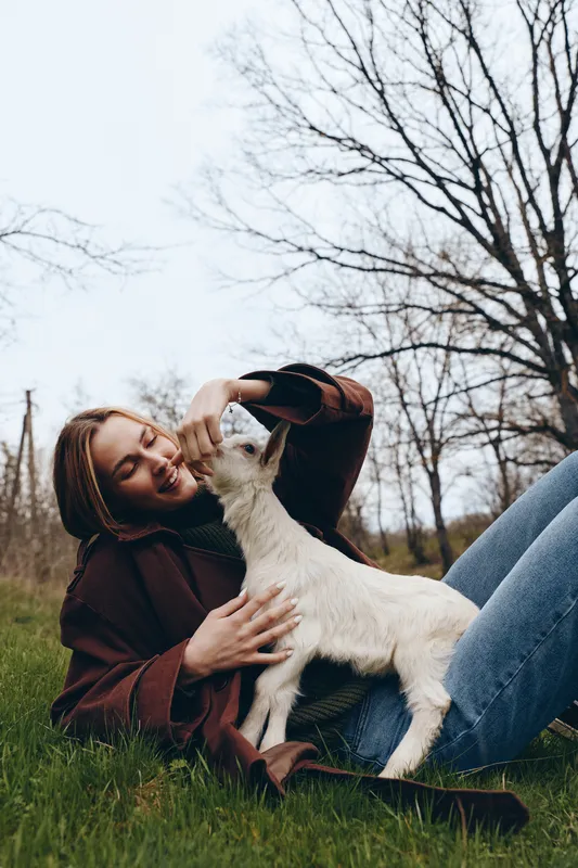 Young woman in a brown jacket lies on the grass smiling while gently feeding a white baby goat in a natural outdoor setting during early spring.