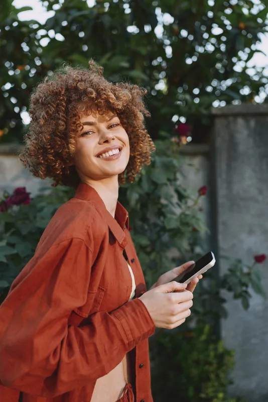 Cheerful woman outdoors, smiling with bright eyes, casual red jacket in a sunlit garden, a relaxed lifestyle moment, phone in hand, natural colors, candid portrait