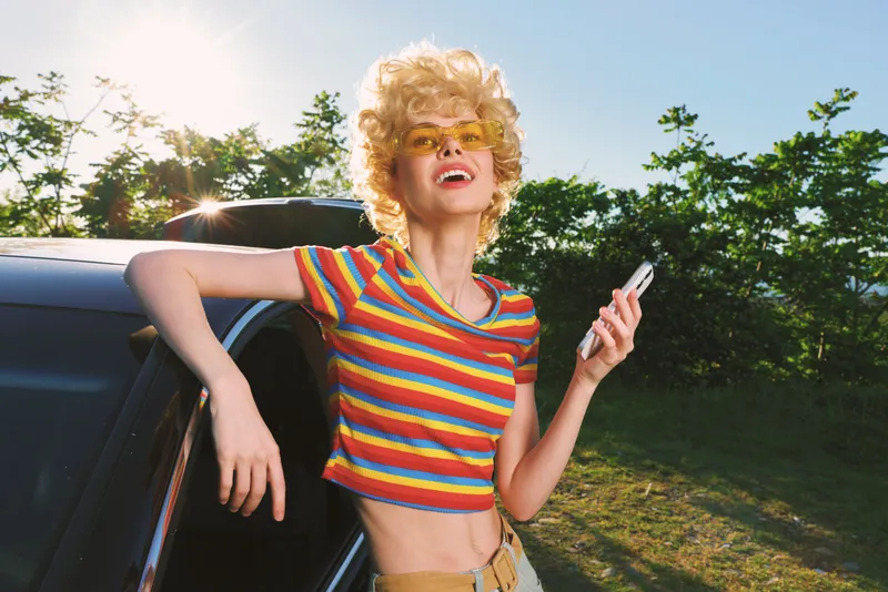 Smiling woman with blonde hair in a rainbow striped crop top leans on a car, sunshine and outdoor setting. Retro style warmth, carefree fashion, summer lifestyle portrait.