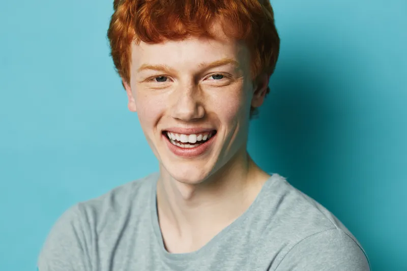 Portrait of smiling redhead man with freckles and casual grey t shirt looking at camera on blue background. Friendly expression with natural light for lifestyle concept.