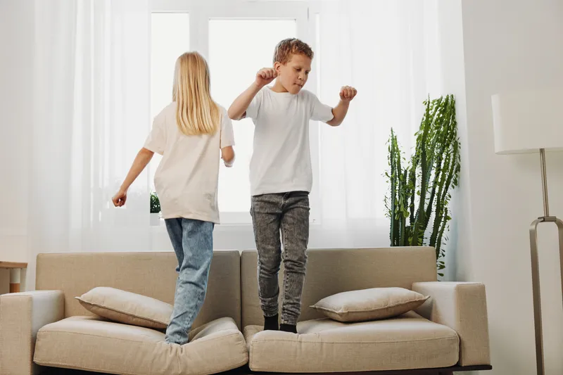 Two children wearing casual clothes joyfully jump on a beige sofa in a bright living room with large windows and green plants. Concept of childhood, play, and home activity.