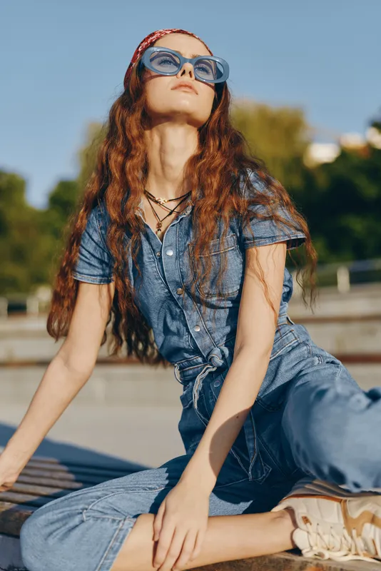 young woman wearing denim outfit and blue sunglasses sitting outdoors on sunny day with confident expression and casual style in summer