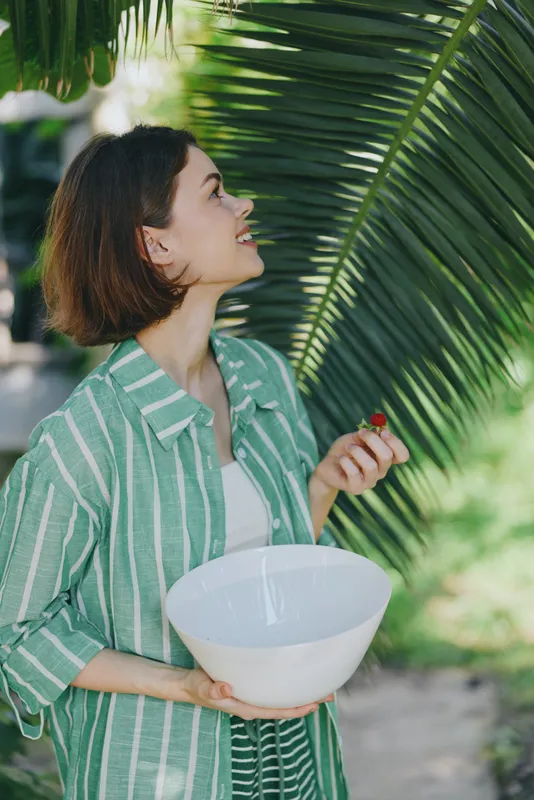 Woman holding a white bowl beneath large palm leaves, outdoor light, casual striped jacket, and a sunny garden mood.