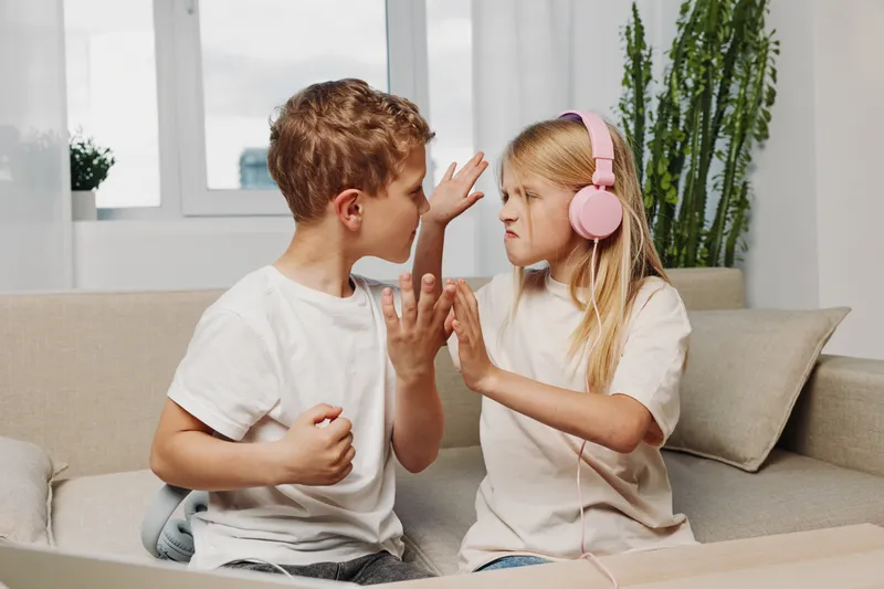Boy and girl quarrel while sitting on couch in living room, girl wearing headphones blocks boy with hand. Concept of sibling conflict, childhood emotions, and family dynamics indoors.