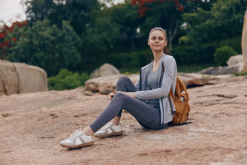 Young woman in activewear sitting on rocky terrain, expressing a relaxed smile, set against a natural green background, ideal for wellness and outdoor lifestyle themes